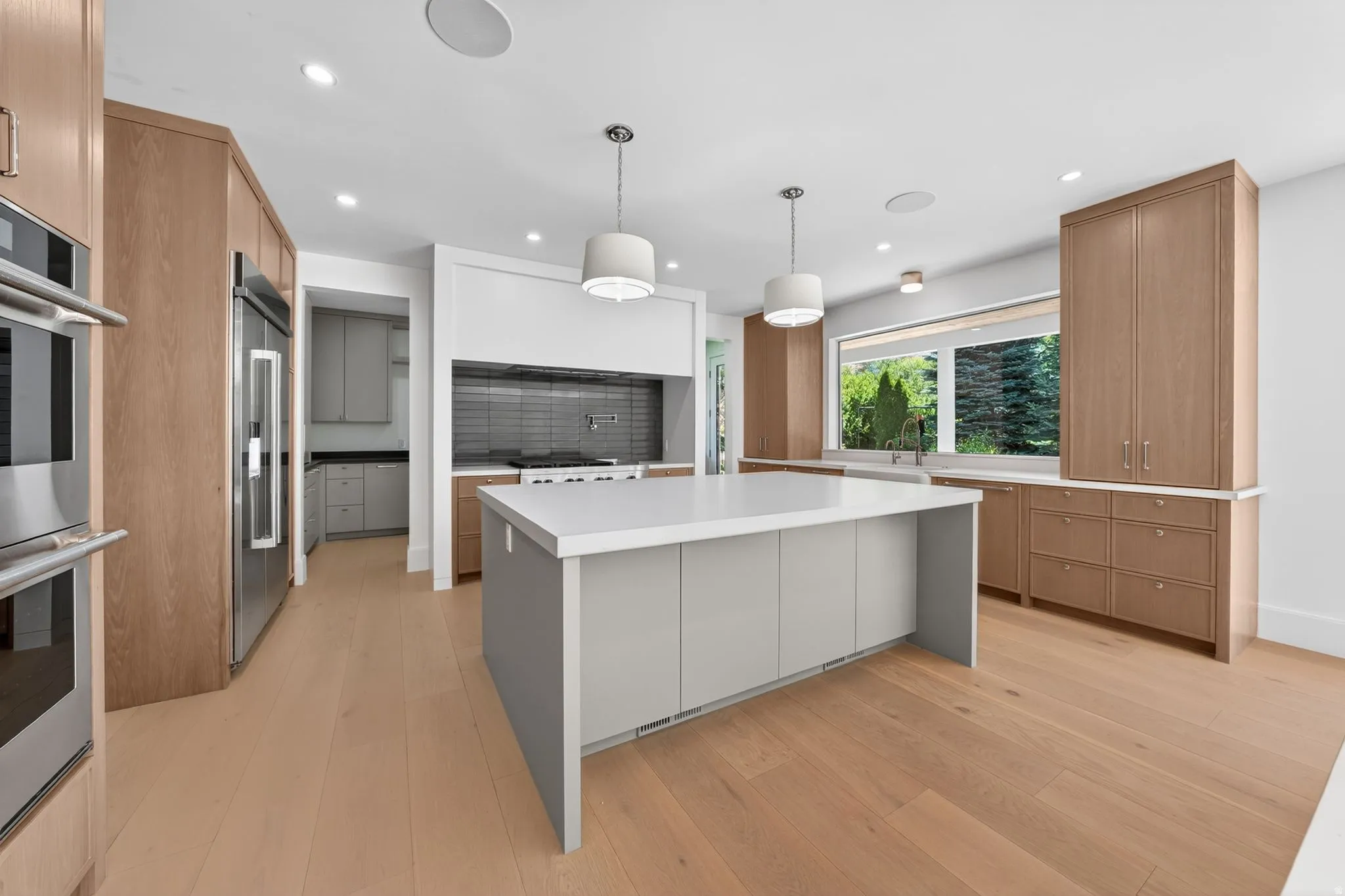 Open-concept kitchen featuring light wood-finish flooring, a central island with a white countertop, recessed lighting, and stainless steel appliances including double ovens and a refrigerator