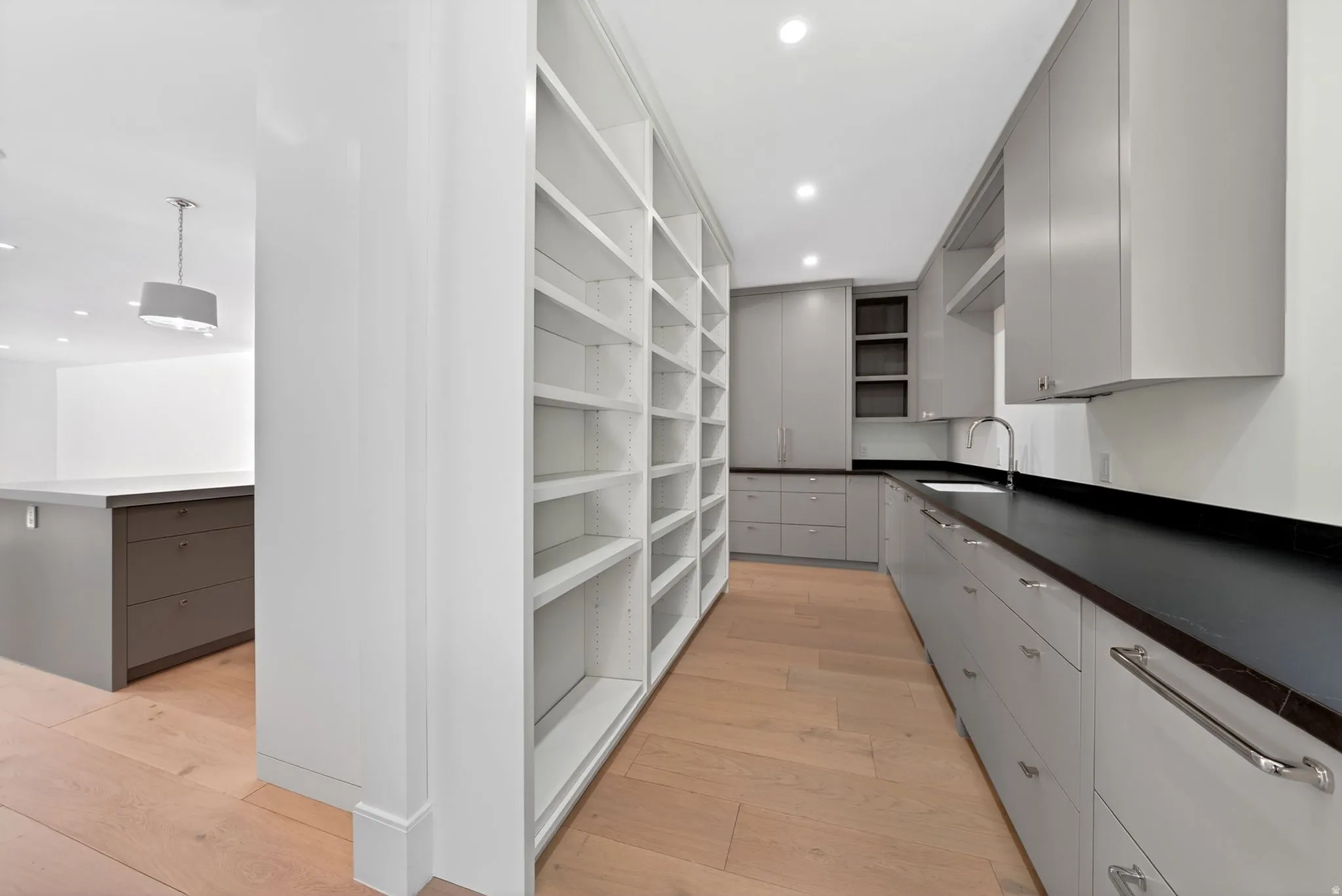 Butler Pantry with spacious kitchen featuring extensive built-in shelving, light wood-finish flooring, recessed lighting, gray cabinetry, and dark countertops with an undermount sink