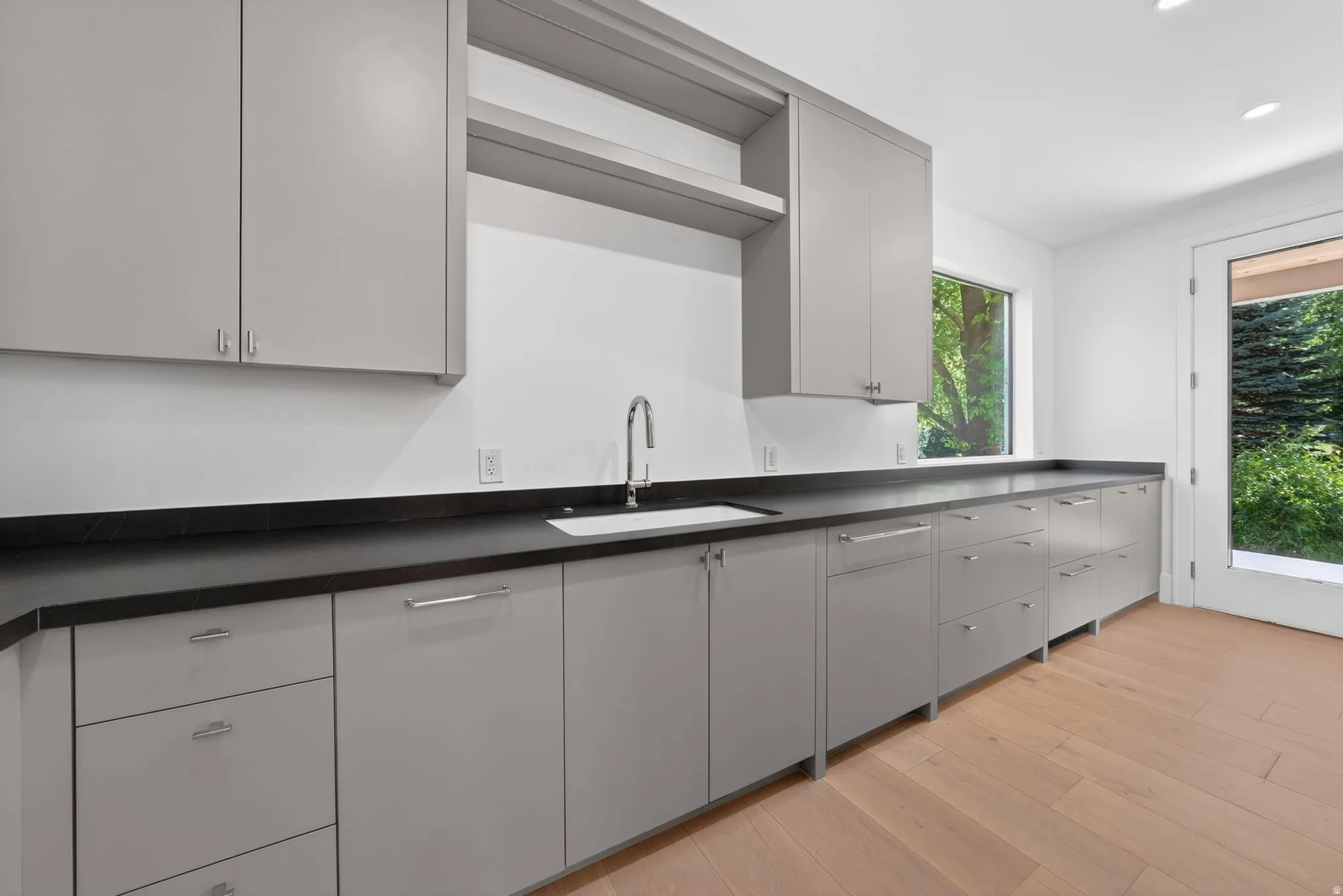Butler pantry with contemporary kitchen featuring light gray cabinetry, dark countertops, a stainless steel sink with a gooseneck faucet, light wood-finish flooring, and a glass-paned door with access to backyard