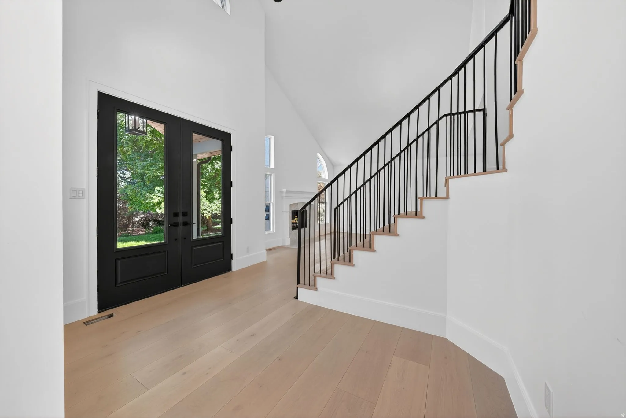Double entry doors with full glass panels and matte black hardware, light wood-finish flooring, white baseboards, and a tall vaulted ceiling