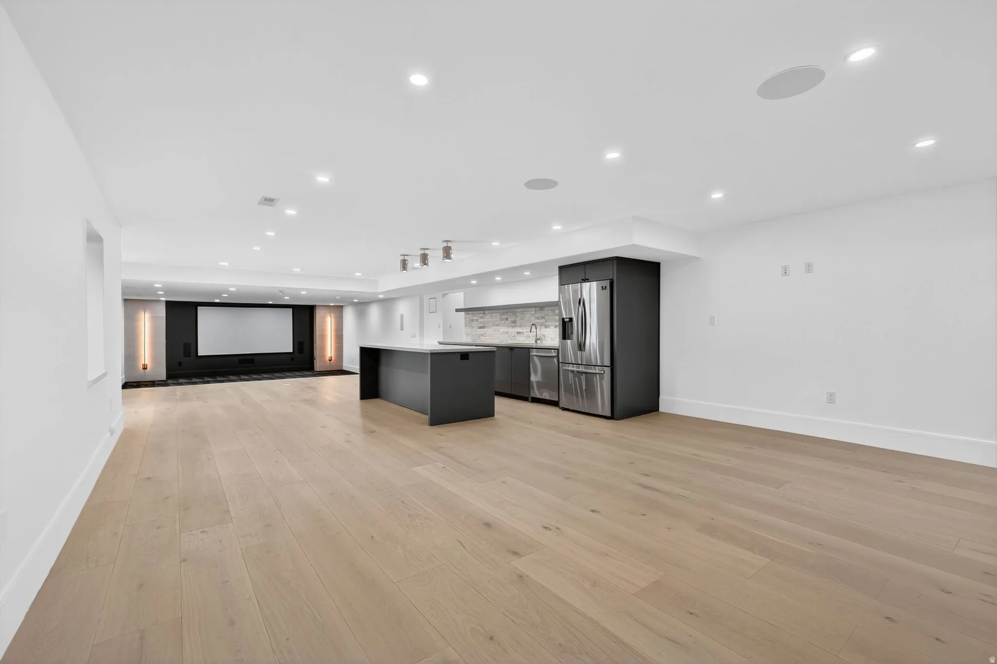 Expansive entertainment space featuring light wood-finish flooring, recessed lighting, and a built-in media wall with projection screen