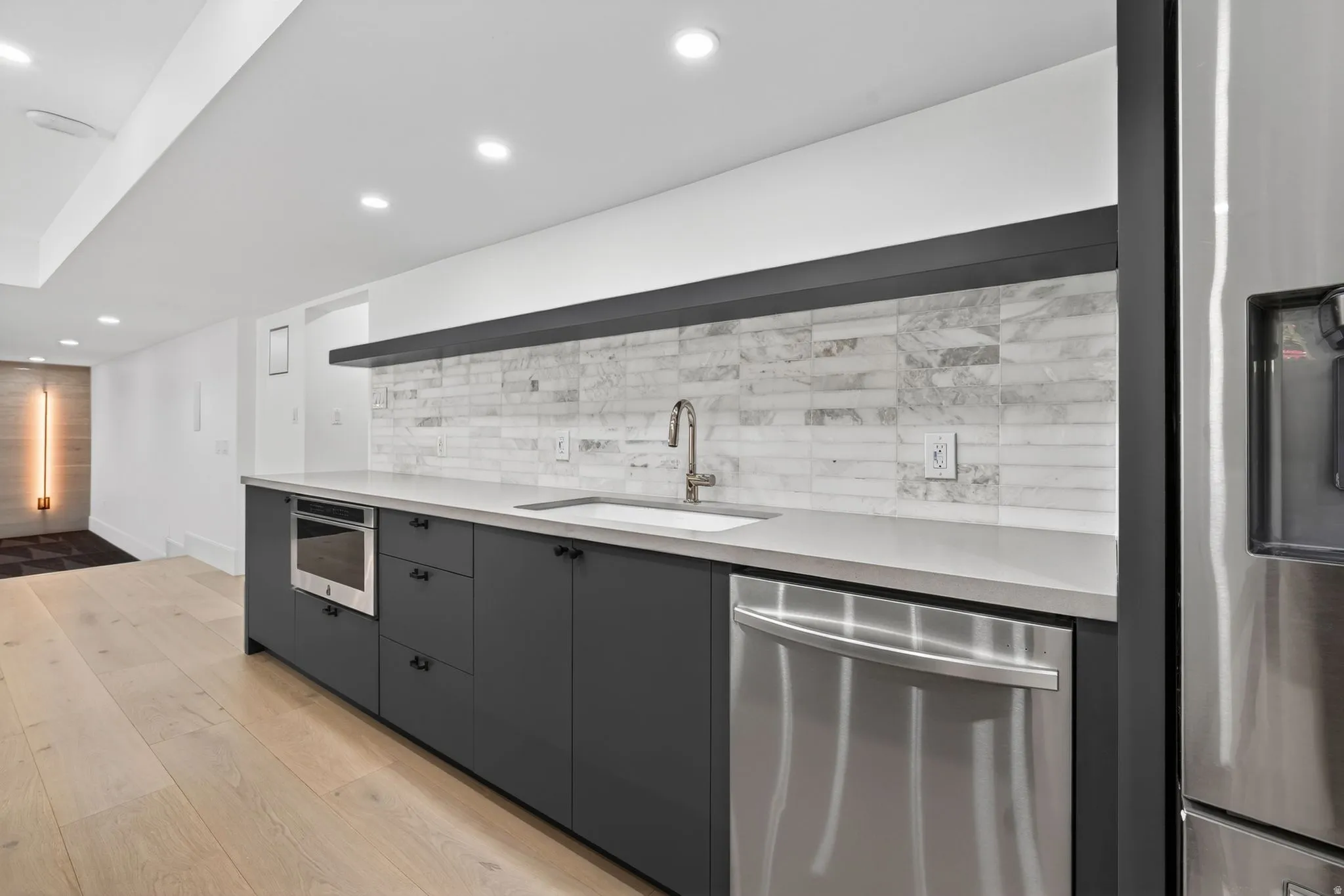 Kitchenette featuring light wood-finish flooring, dark cabinetry, light countertops, a white subway tile backsplash, and stainless steel appliances including a built-in microwave and dishwasher