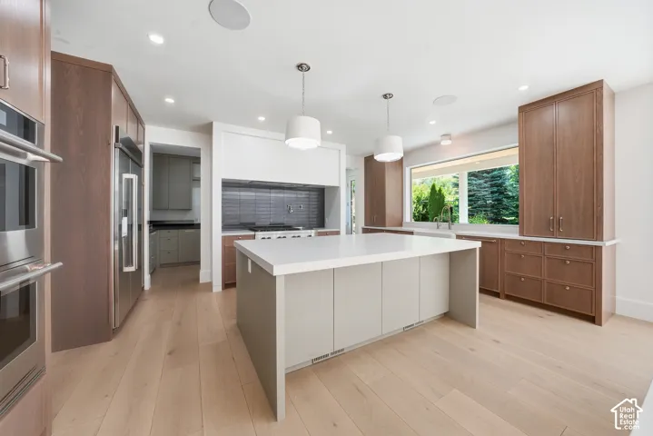 Spacious kitchen featuring a large central island with a white countertop, light wood-finish flooring, integrated stainless steel appliances, extensive cabinetry with light brown wood finish, and a wide horizontal window with exterior landscaping