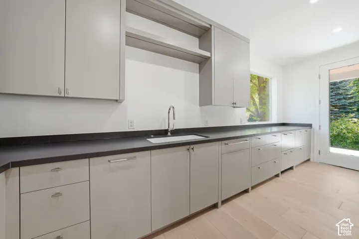 Modern butler pantry featuring extensive light grey cabinetry with chrome hardware, window outlooking to back yard, dark countertops, a stainless steel sink with a gooseneck faucet, light wood-finish flooring, and a glass-paned exterior door with access t