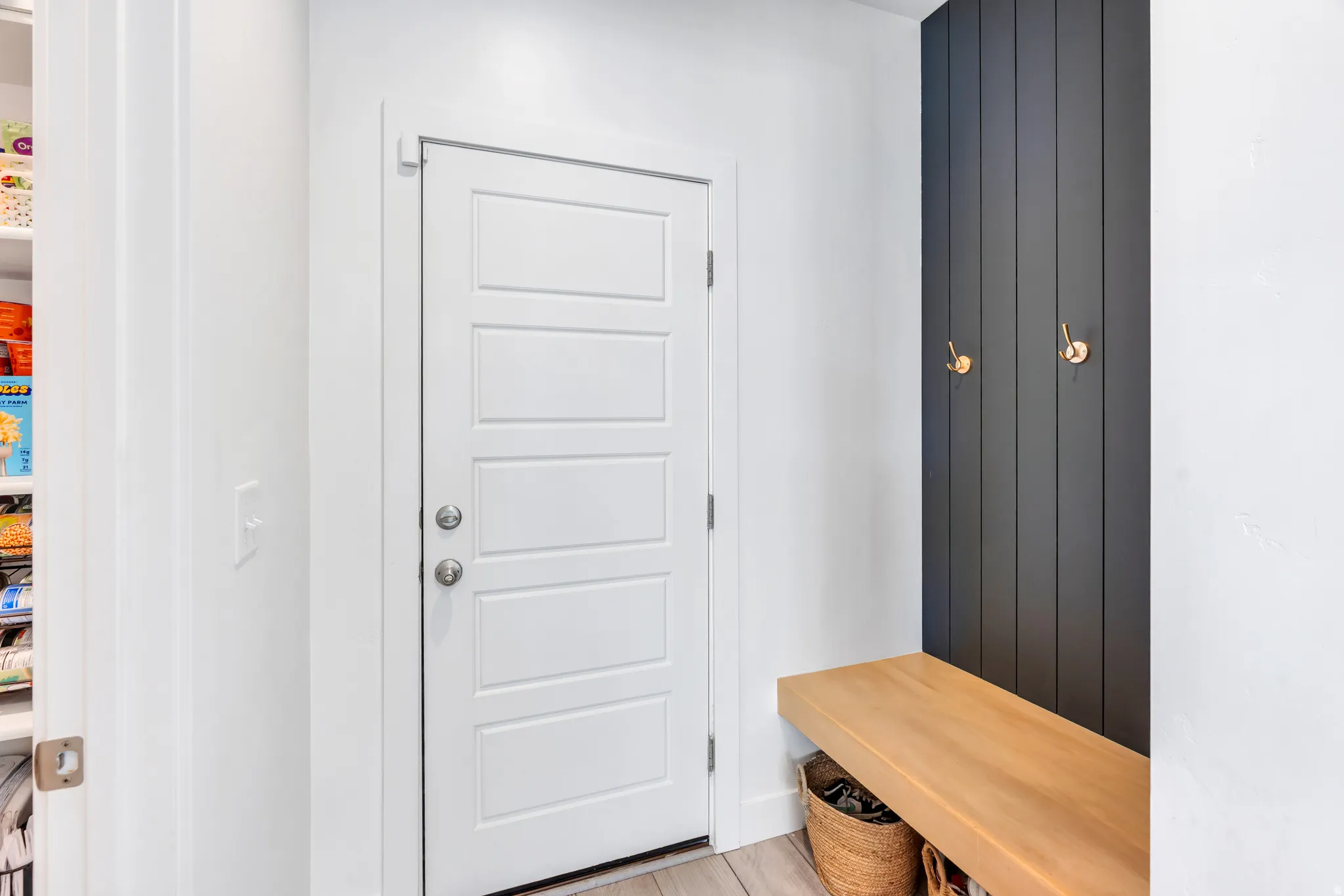 Entryway mudroom area featuring a white paneled door, a built-in wooden bench, and a dark vertical plank wall with two brass hooks