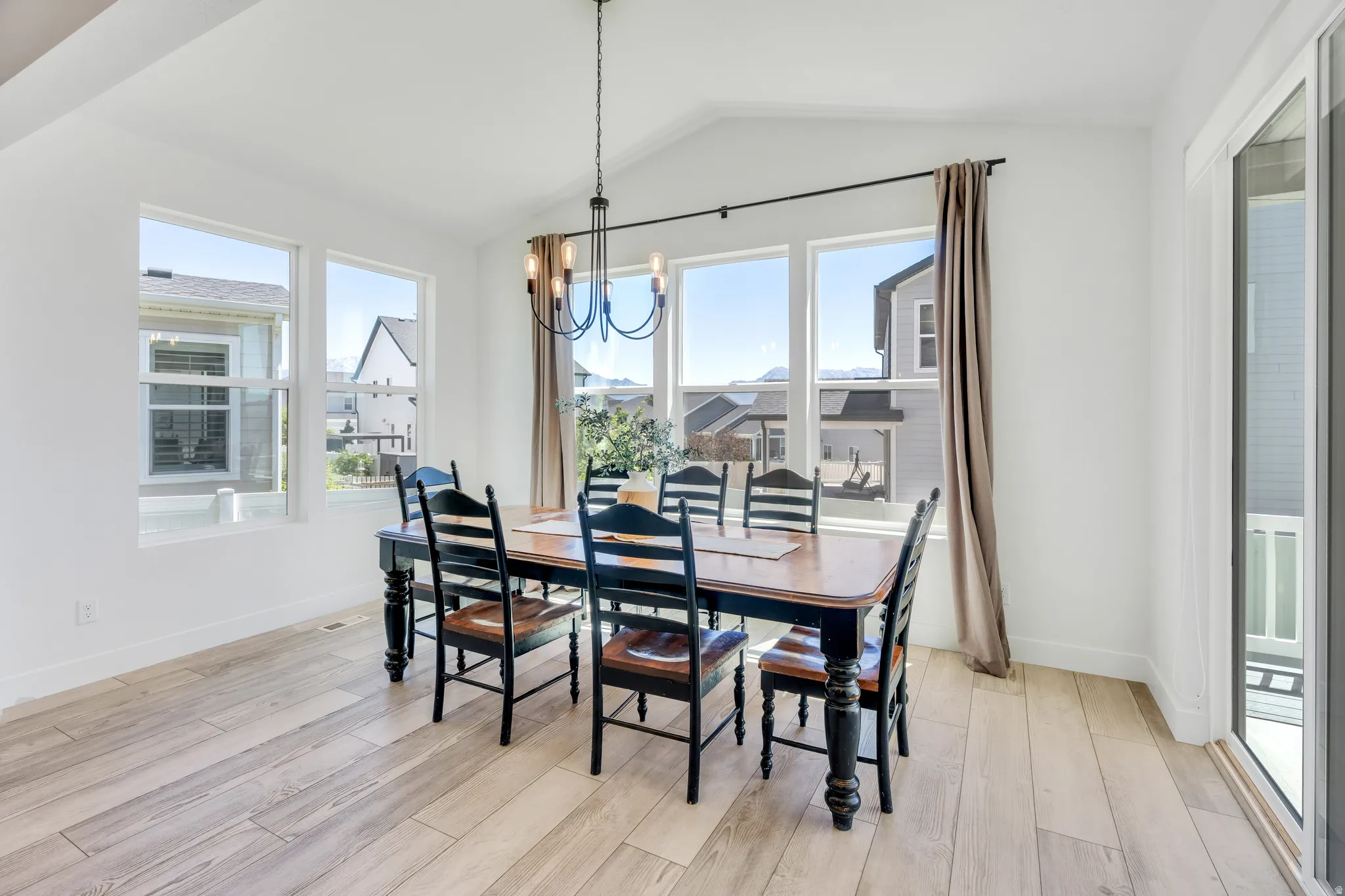 Bright dining area featuring vaulted ceilings, light wood-finish flooring, multiple large windows, a contemporary chandelier, and a glass sliding door