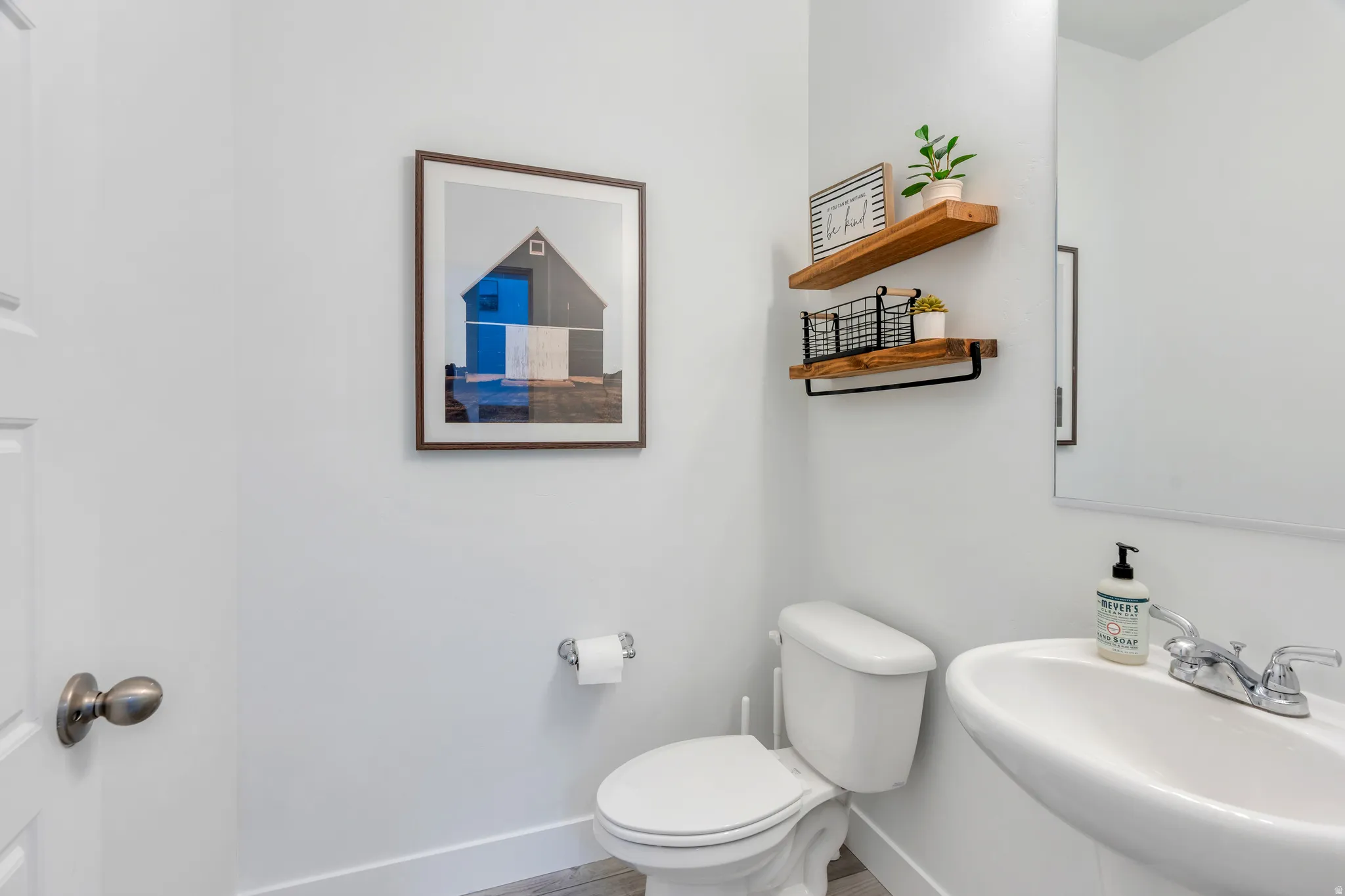 Powder room featuring a pedestal sink with chrome fixtures, a two-piece toilet, wood-finish floating shelves, and light-colored flooring
