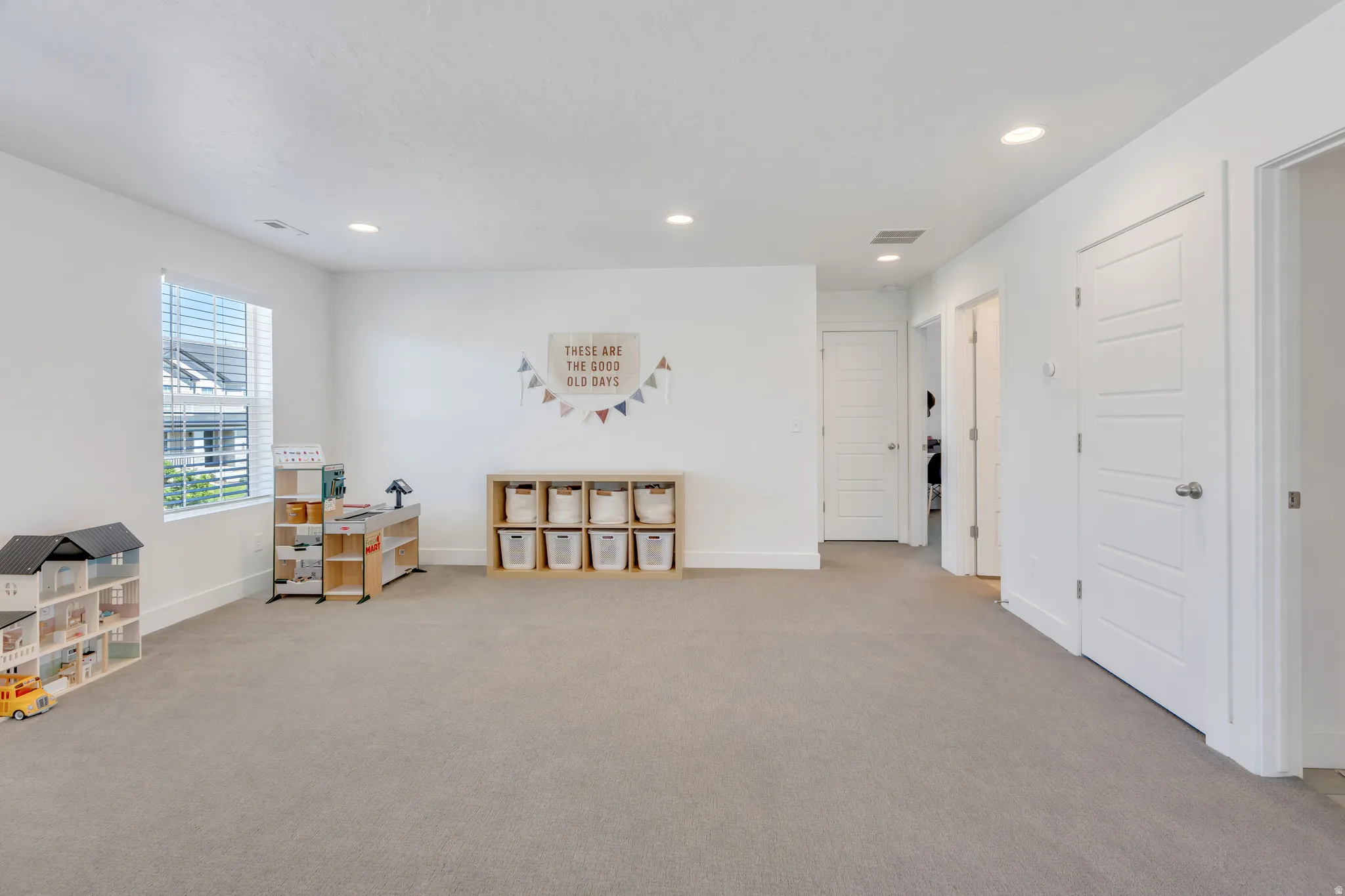 Spacious room featuring light gray carpeting, crisp white walls, recessed ceiling lighting, a window with horizontal blinds, and multiple white paneled doors