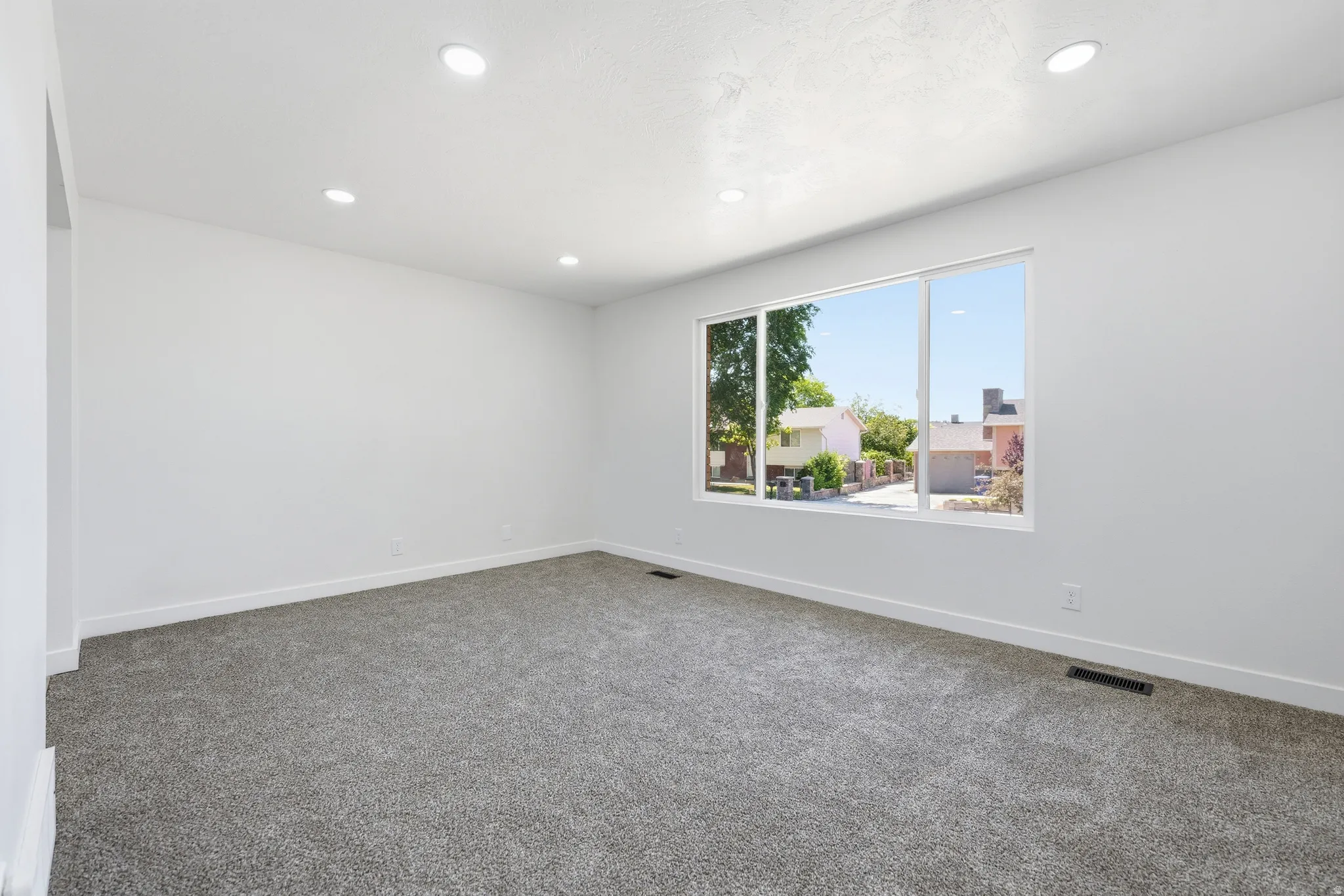Spacious interior room featuring light gray carpeting, recessed ceiling lighting, a large window, white baseboards, and a neutral wall color