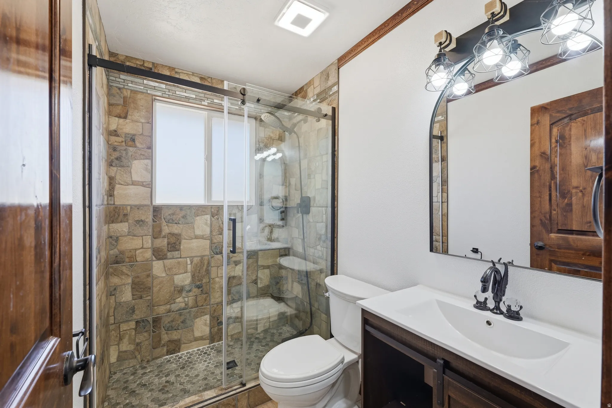 Bathroom featuring a stone-tiled shower with glass enclosure, single vanity with integrated sink, dark wood cabinetry, and an arched mirror with industrial-style lighting