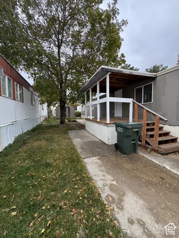 Covered wooden porch with railings, concrete pathway, and detached storage structure situated on a grass lot with mature trees