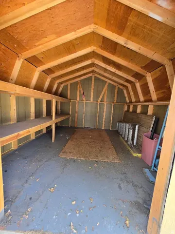 Spacious interior storage shed featuring a gambrel roof design, exposed wood framing, built-in shelving, and painted flooring