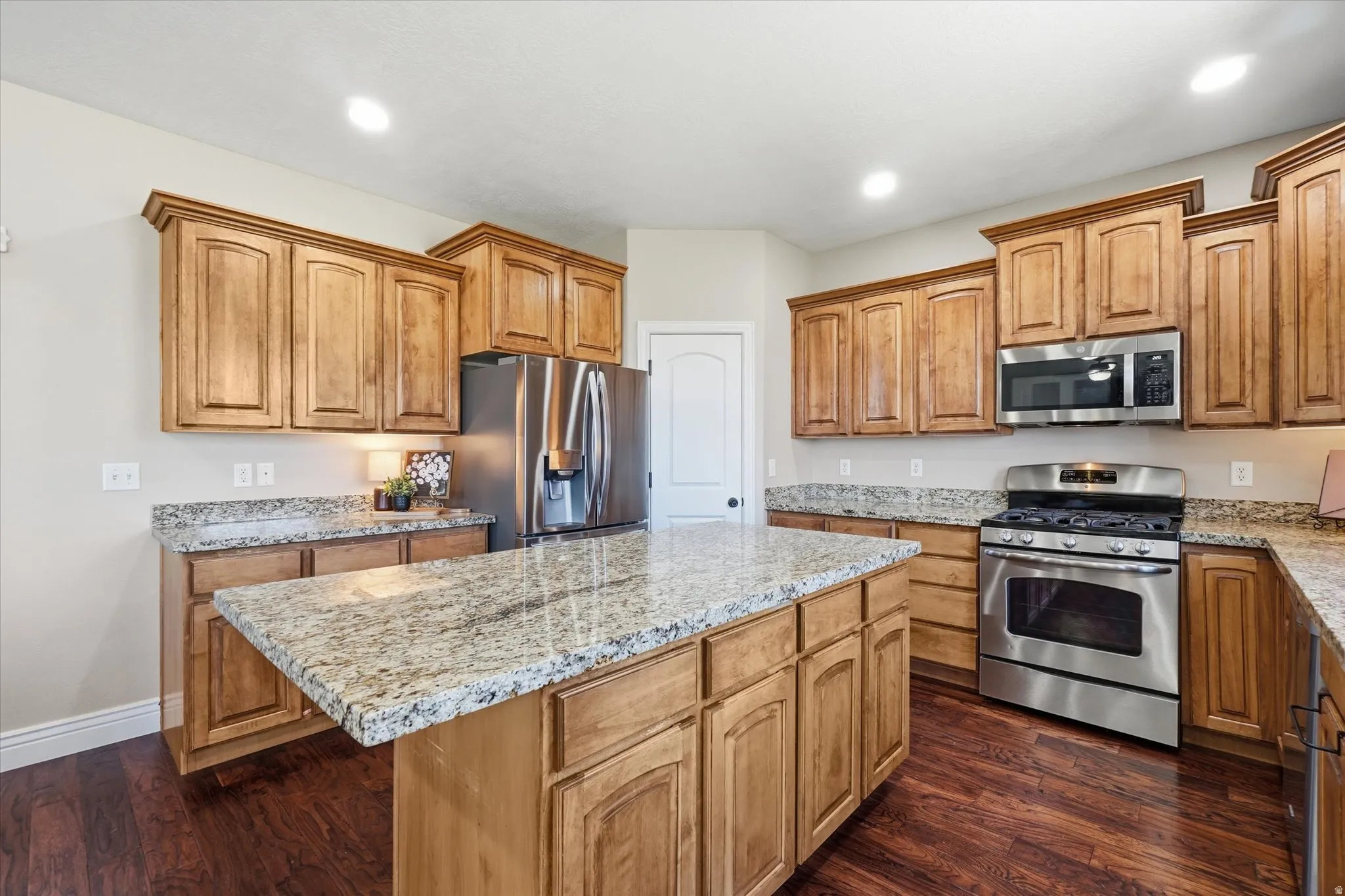 Spacious kitchen featuring a large central island with light granite countertops, extensive cabinetry, stainless steel appliances, and wood-finish flooring