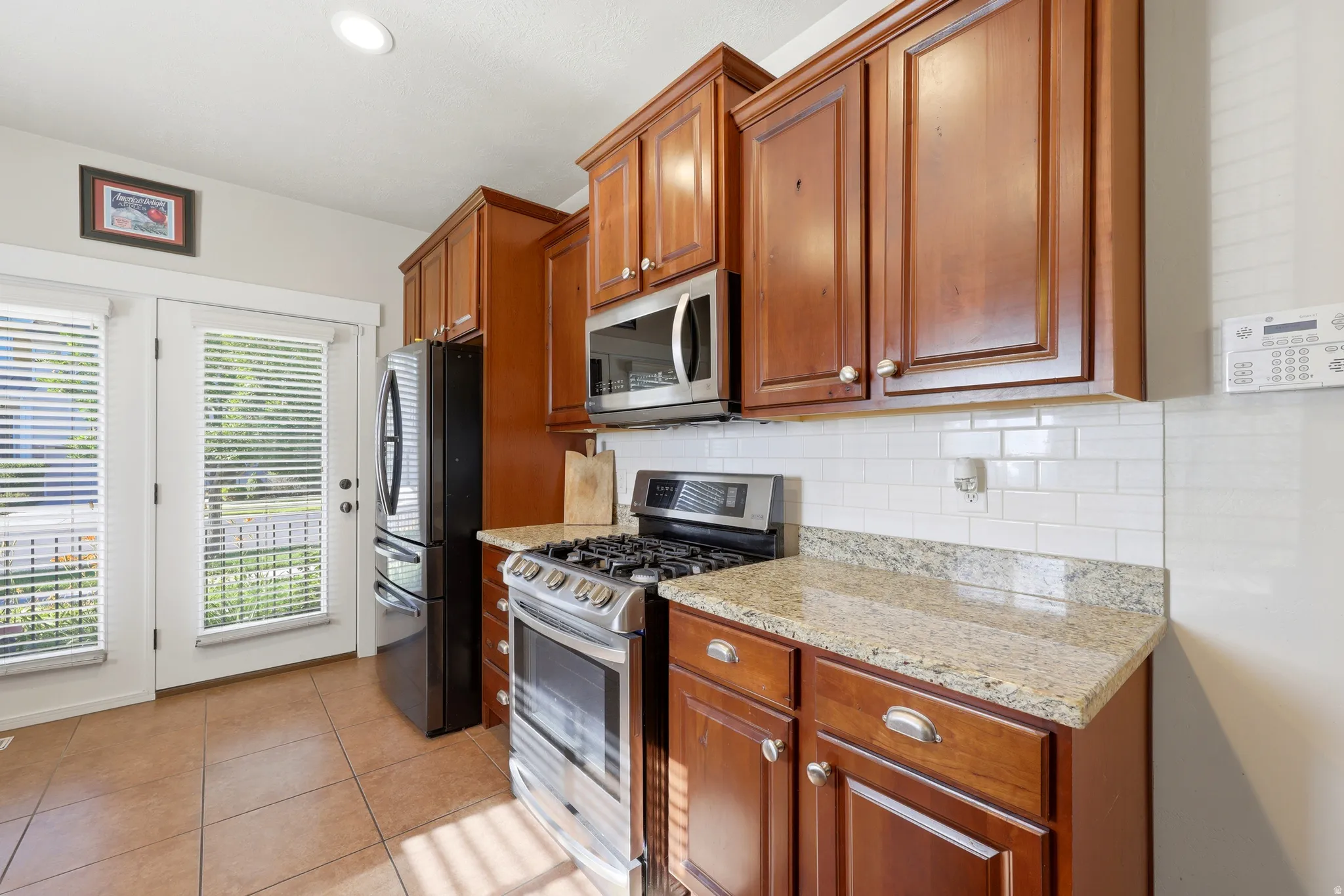 Well-appointed kitchen featuring wood cabinetry, granite-finish countertops, stainless steel appliances, a subway tile backsplash, and tile flooring