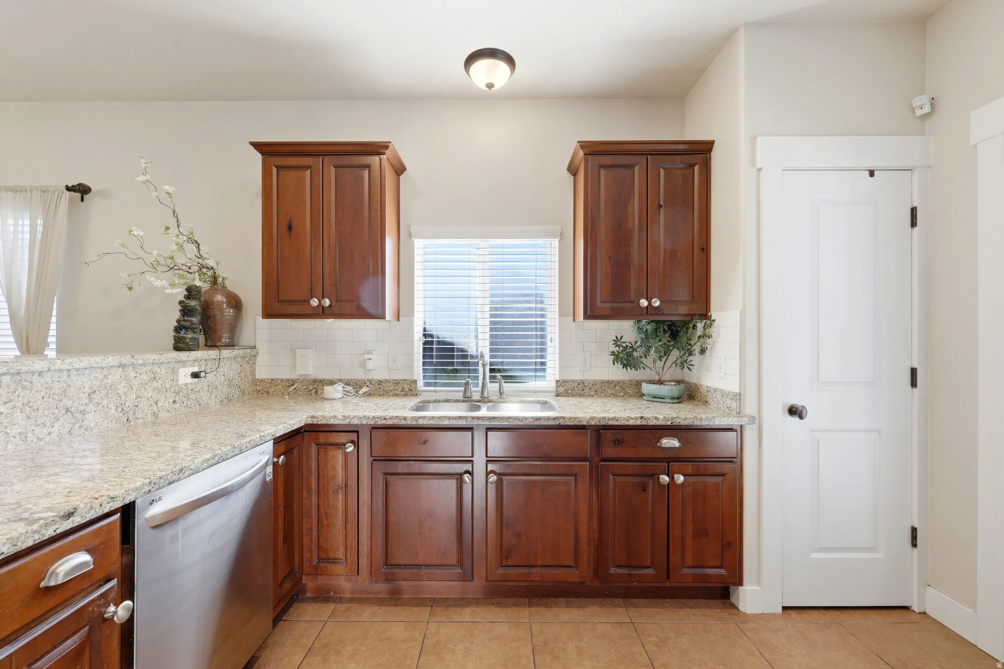 Kitchen featuring wood cabinetry, light-toned countertops, a white tile backsplash, a stainless steel dishwasher, and a window with blinds