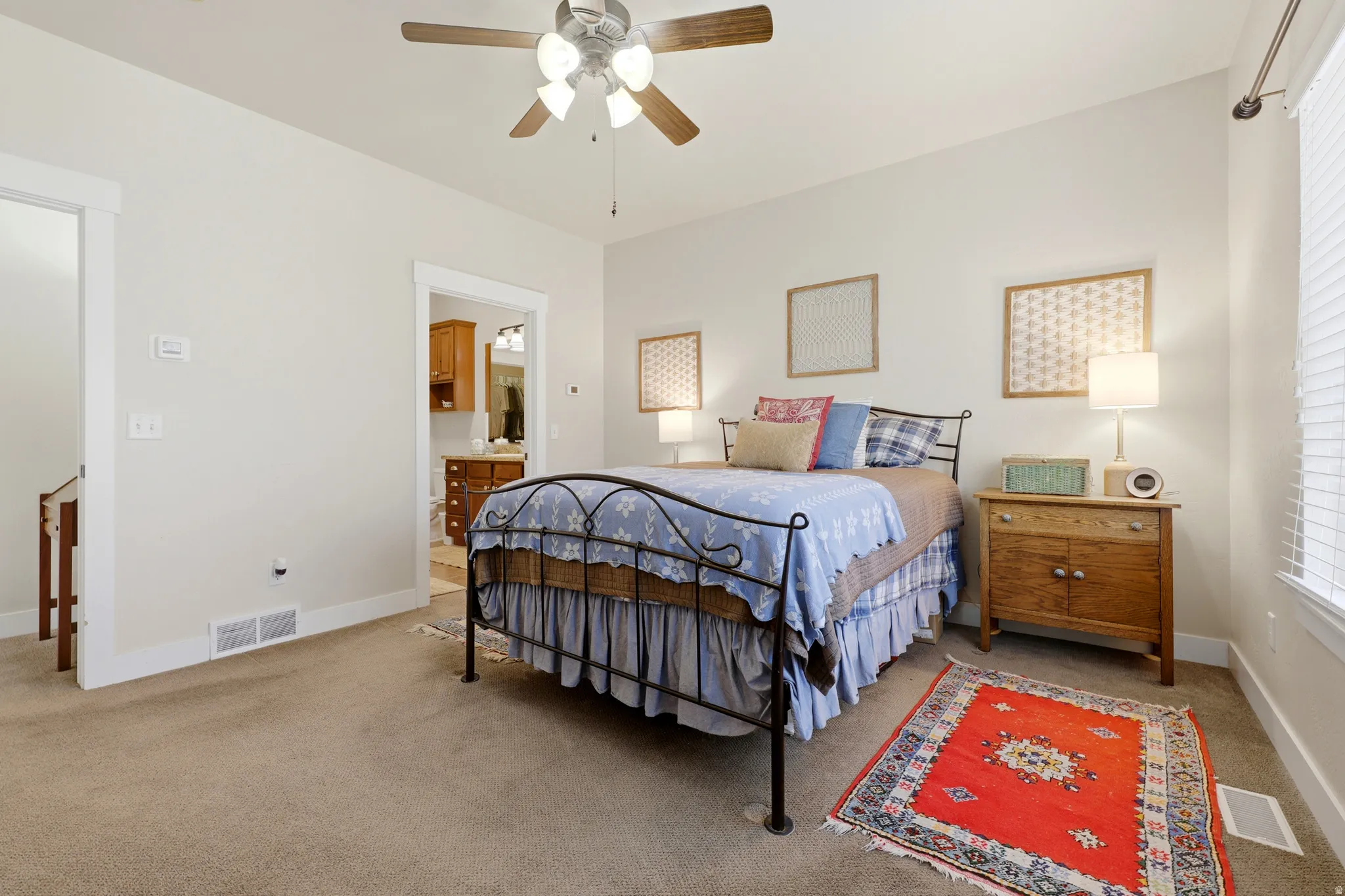 Carpeted bedroom featuring a ceiling fan with light fixture, window with blinds, and an open doorway to a bathroom with wood cabinetry
