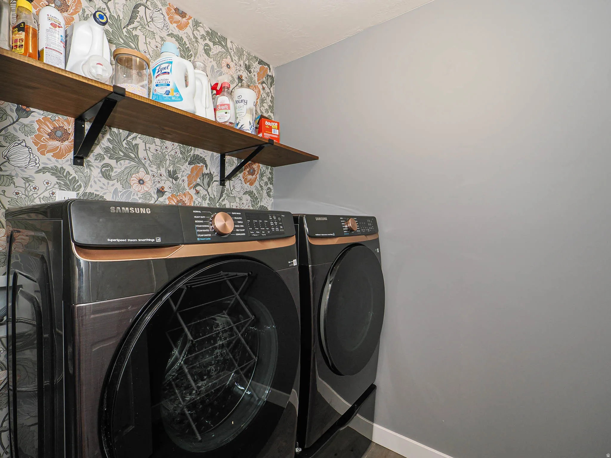 Dedicated laundry space featuring a wood-finish floating shelf, decorative botanical wallpaper, and neutral gray painted walls