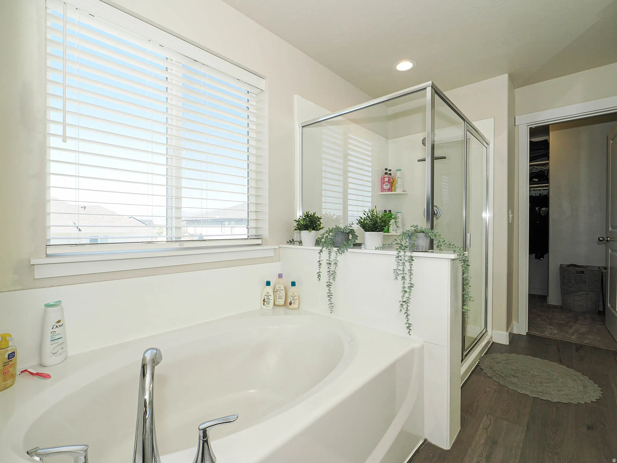 White soaking tub with chrome fixtures, glass-enclosed shower, white window blinds, recessed lighting, and wood-finish flooring