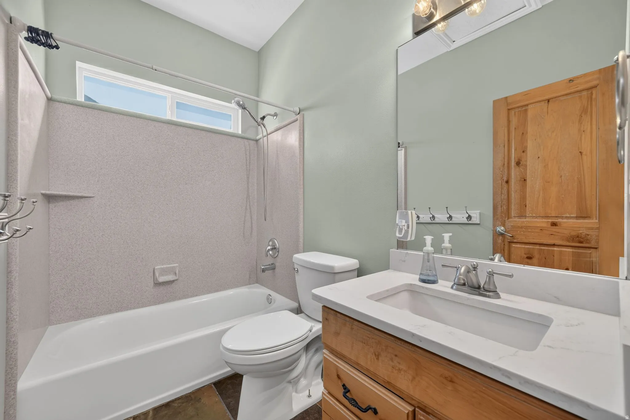 Modern bathroom featuring a white bathtub with a speckled surround, a large vanity with a white countertop and integrated sink, a light wood-finish vanity cabinet, a wall-mounted mirror, and a wood-paneled door