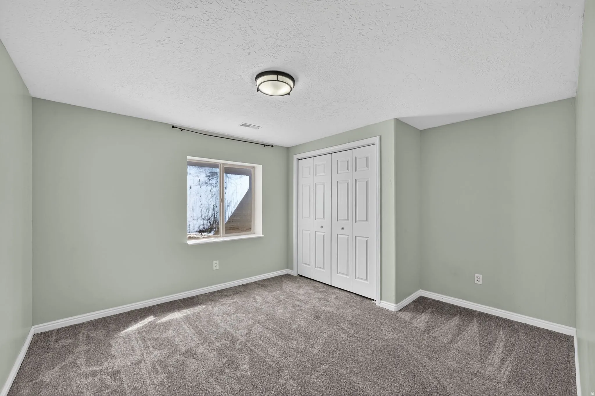 Carpeted room featuring a window, bi-fold closet doors, a ceiling-mounted light fixture, and light green walls with white trim