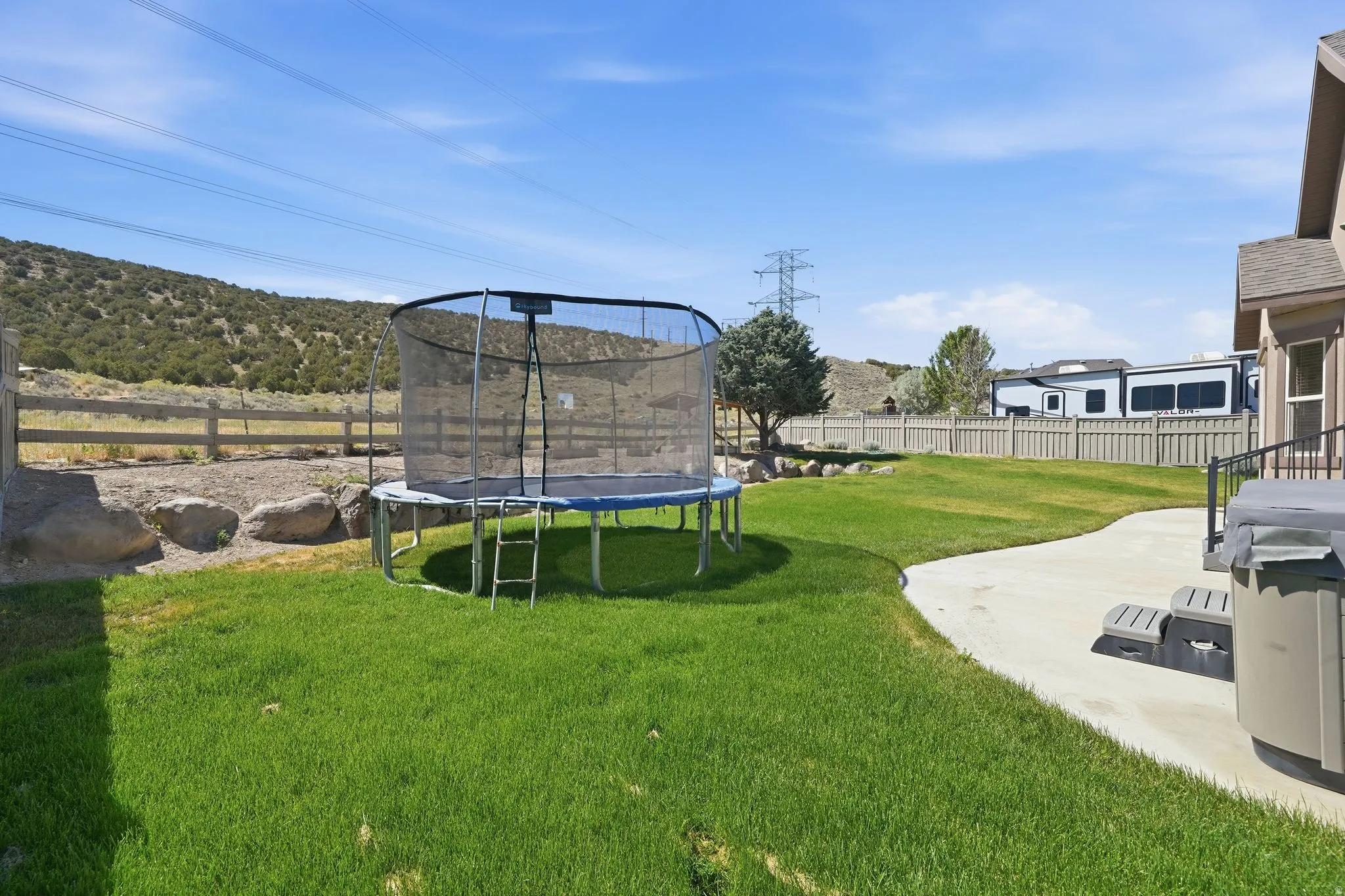 Expansive grassy backyard featuring a curved concrete patio, a built-in spa, a privacy fence, and distant natural landscaping