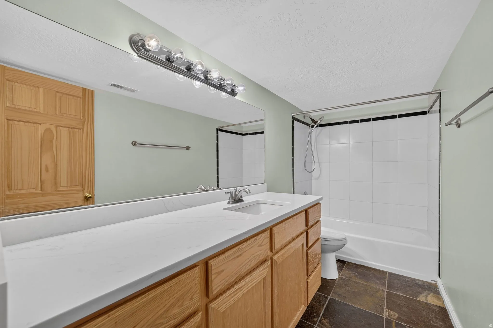 Bathroom featuring a wood vanity with an integrated sink, a large mirror, a chrome light fixture, a white tiled shower/tub combination with black trim, and natural stone flooring