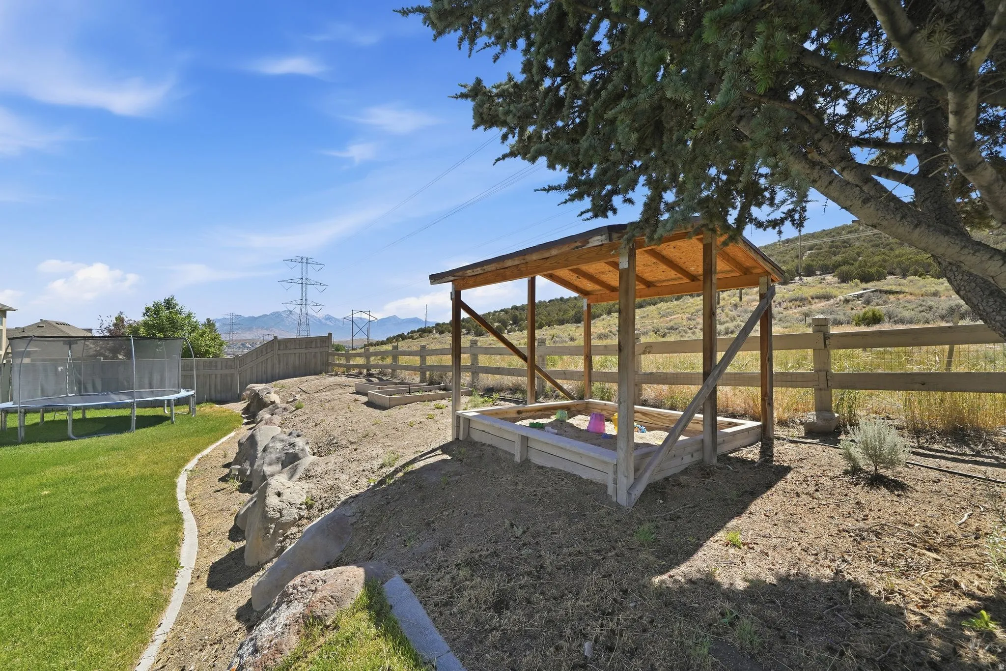Expansive backyard featuring a natural rock retaining wall, a dedicated sandbox area with a wooden shade structure, a manicured lawn, and a wooden perimeter fence