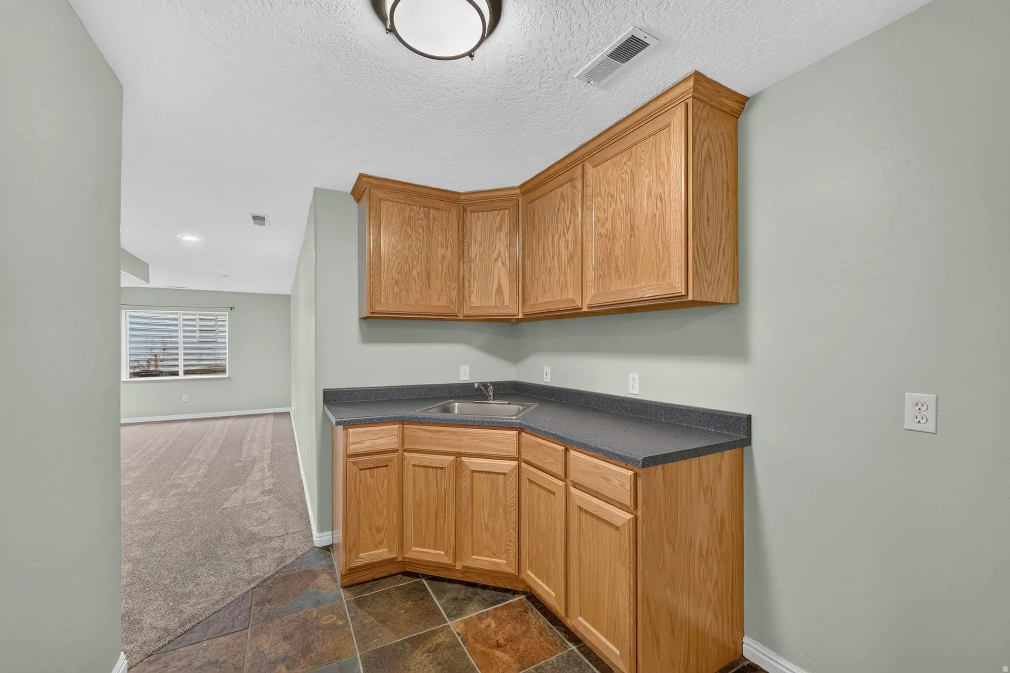 Corner wet bar featuring light wood cabinetry, a dark countertop, and an integrated stainless steel sink