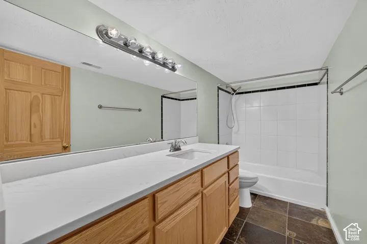 Bathroom featuring a wood vanity with an integrated sink, a large mirror, a chrome light fixture, a white tiled shower/tub combination with black trim, and natural stone flooring