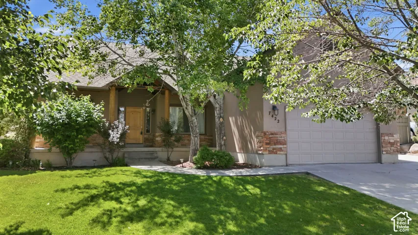 Ranch-style exterior featuring a neutral stucco finish, stone veneer accents, an attached garage, mature trees, and a concrete driveway