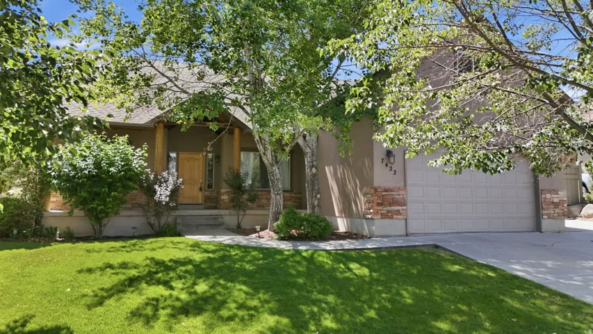 Ranch-style exterior featuring a neutral stucco finish, stone veneer accents, an attached garage, mature trees, and a concrete driveway