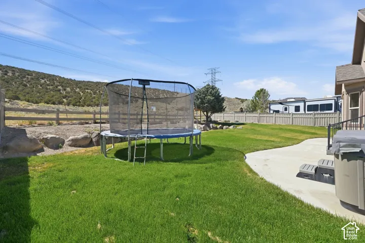 Expansive grassy backyard featuring a curved concrete patio, a built-in spa, a privacy fence, and distant natural landscaping