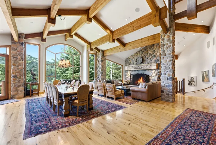 Vaulted ceilings with exposed wood beams, a stone fireplace, and wide-plank wood flooring complement the large arched windows