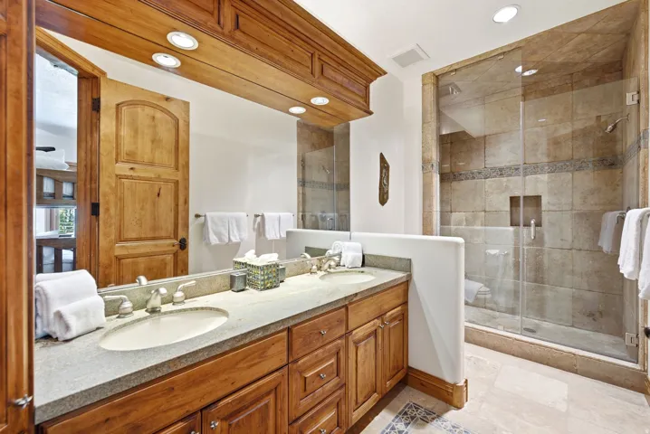 Double vanity with undermount sinks, stone countertop, wood cabinetry, and a glass-enclosed shower with stone tile
