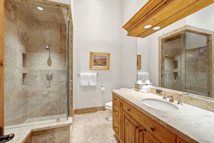 Bathroom featuring a glass-enclosed shower with stone tile surround, a single vanity with wood cabinetry and stone countertop, a large wall-mounted mirror, and stone tile flooring