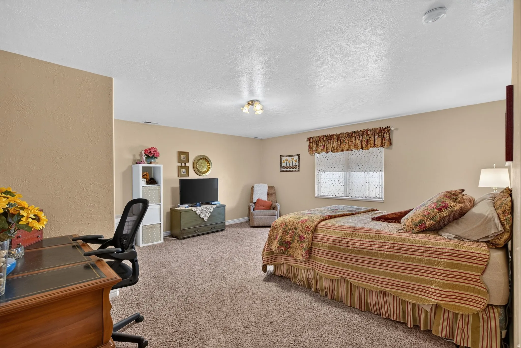 Spacious carpeted room featuring a ceiling-mounted light fixture, light-toned wall paint, a window with a valance, and a visible smoke detector