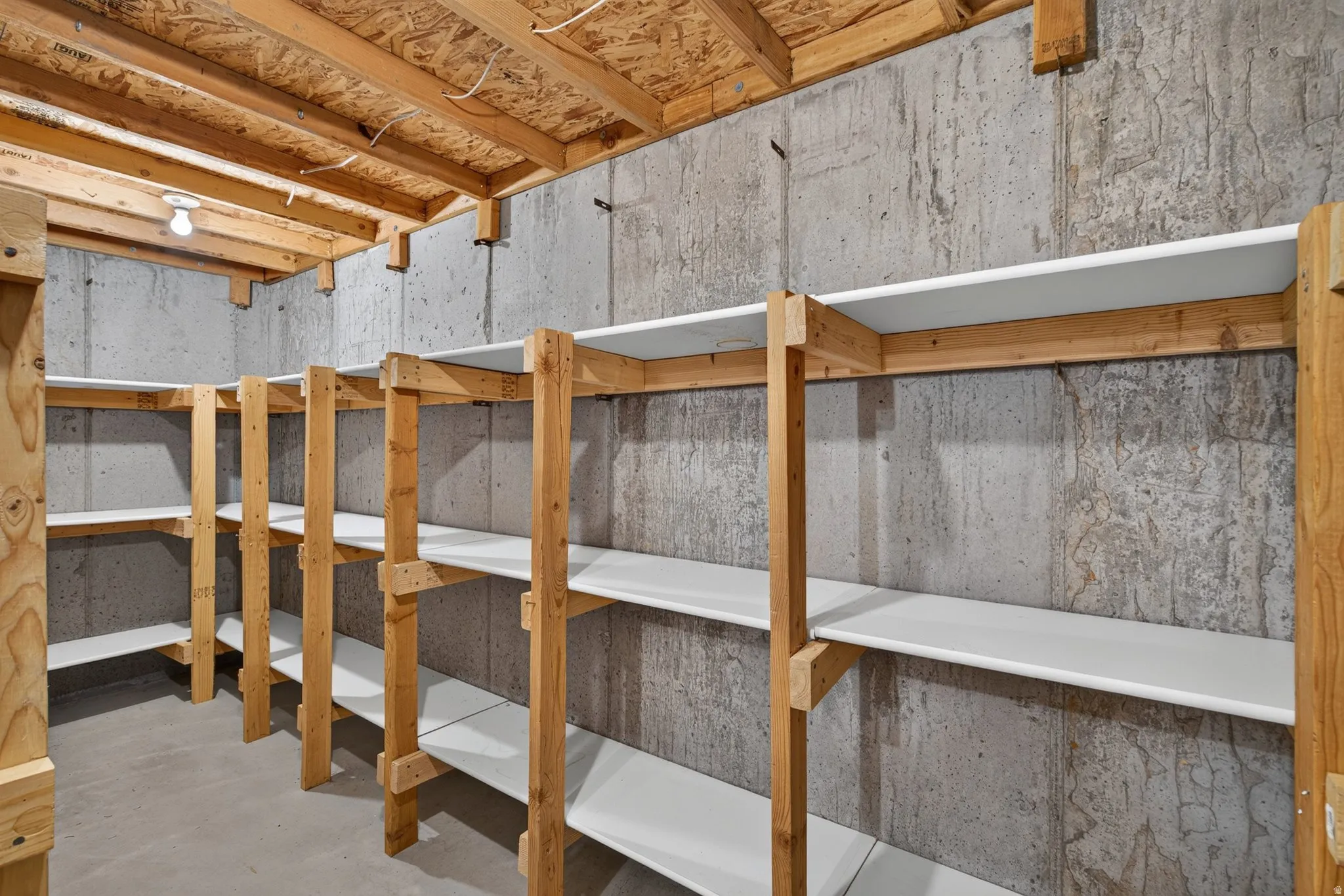 Basement storage room featuring extensive built-in shelving, a concrete floor, concrete walls, and an exposed wood joist ceiling