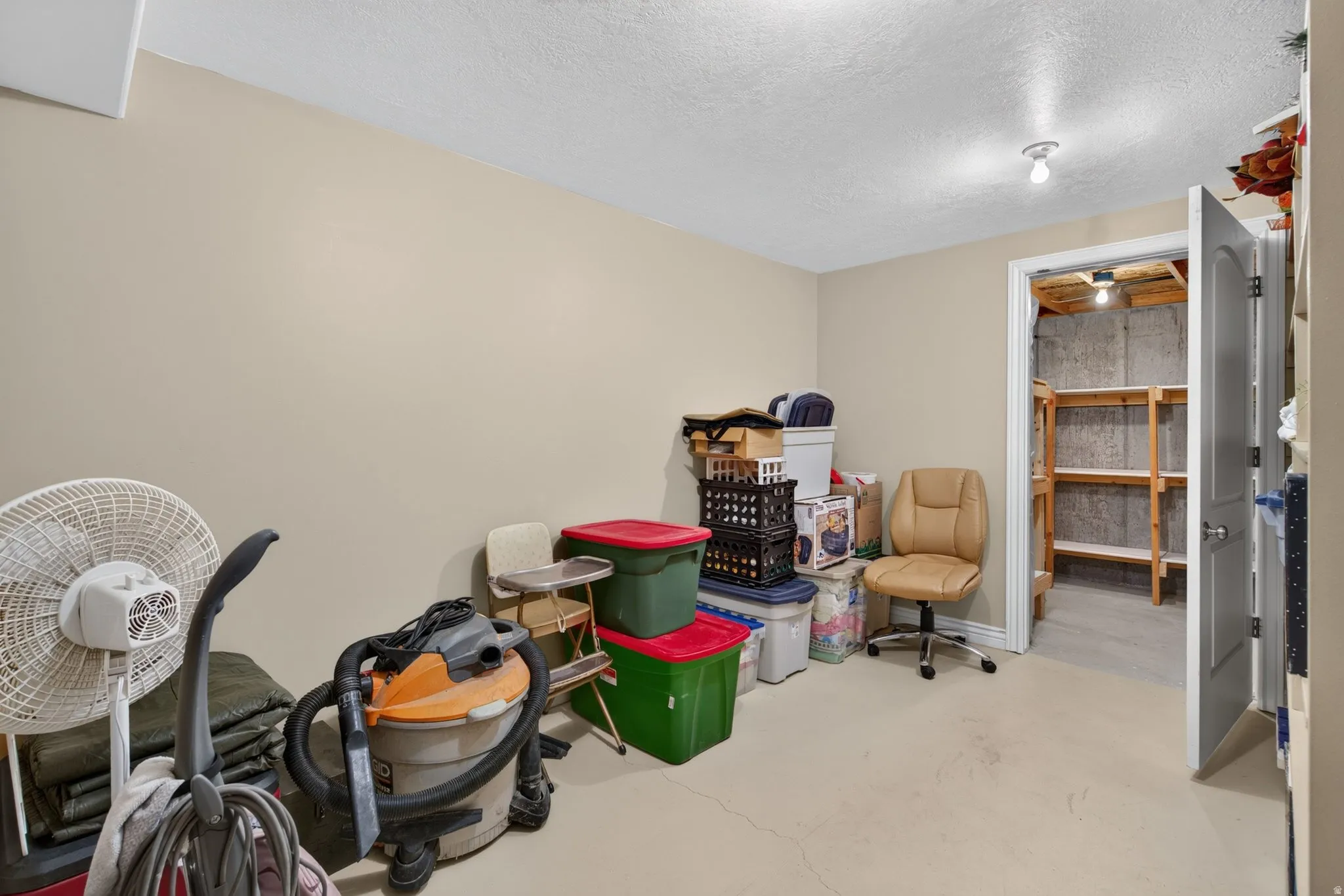 Finished basement room with neutral wall paint and recessed lighting, featuring an open doorway to an unfinished storage area with wood shelving
