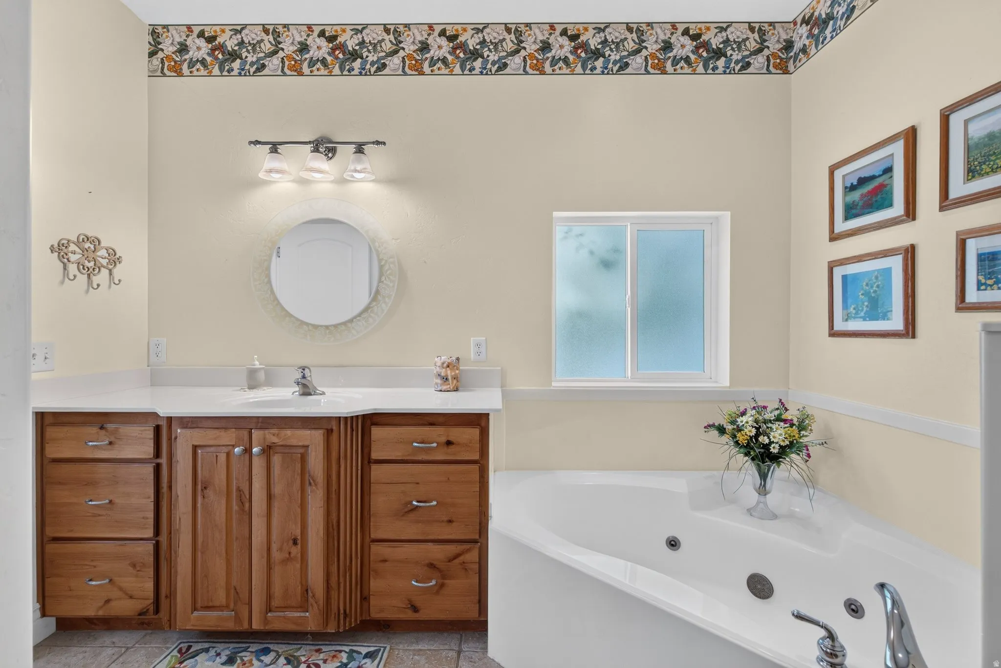 Bathroom featuring a white jetted tub, wood-finish vanity with white countertop, round mirror, and a window with privacy glass