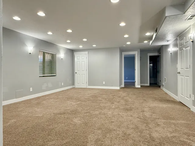 Spacious carpeted room featuring recessed lighting, wall sconces, a window with blinds, and multiple white paneled doors