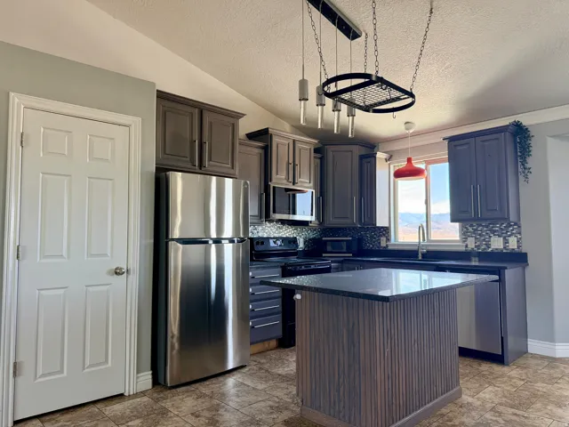 Kitchen featuring dark wood-finish cabinetry, stainless steel appliances, a textured tile backsplash, and an island with a dark countertop