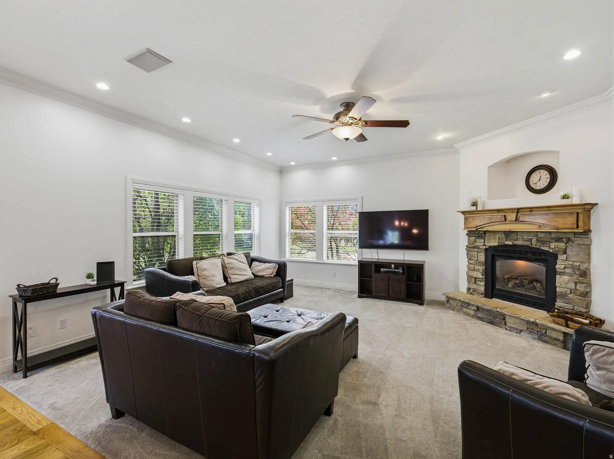 Living area featuring a stone fireplace with a wood mantel, crown molding, recessed lighting, a ceiling fan, and multiple windows