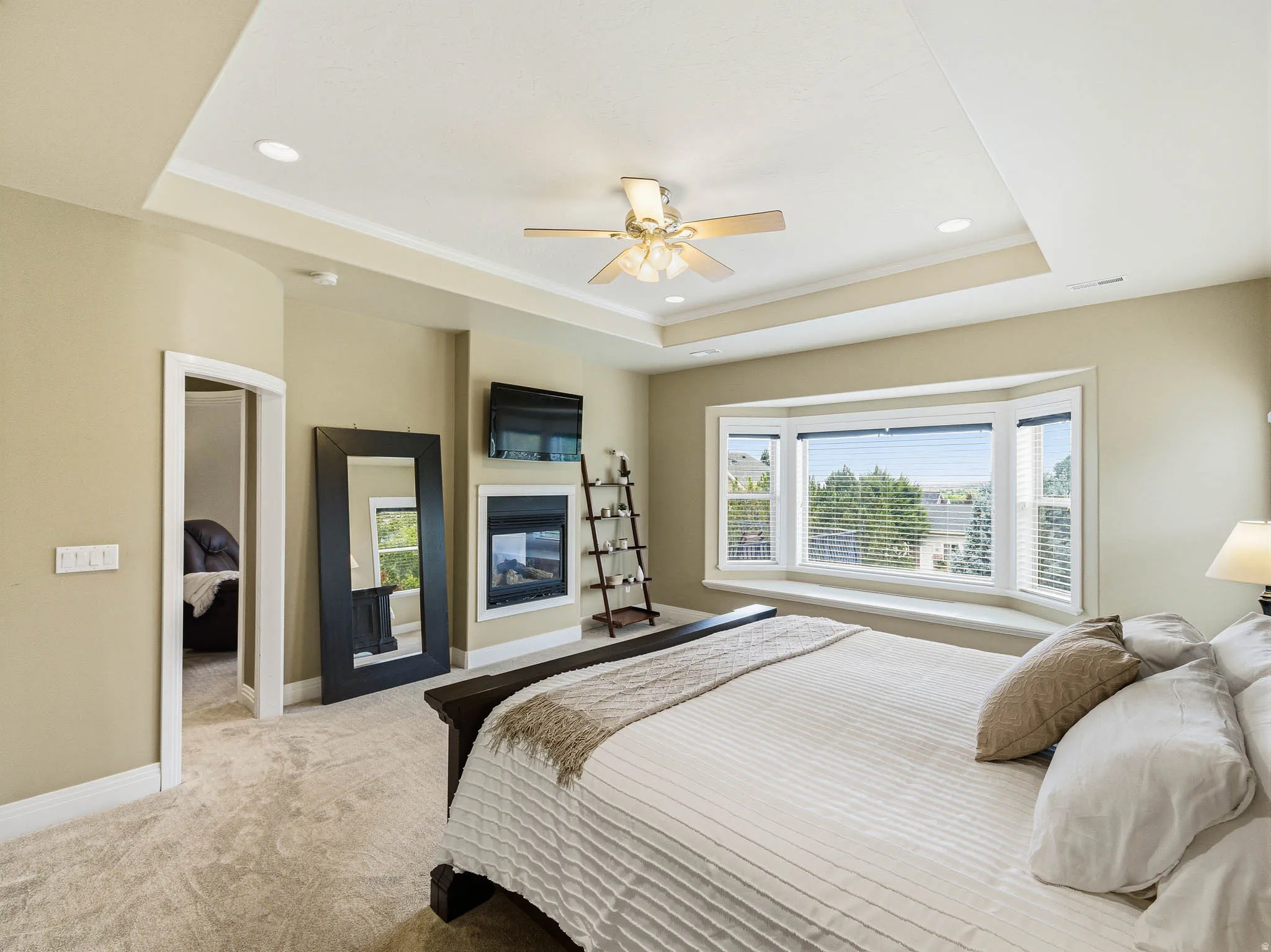 Spacious room featuring a tiered tray ceiling with recessed lighting, a ceiling fan, plush carpeting, a built-in fireplace, and a large bay window with a deep sill