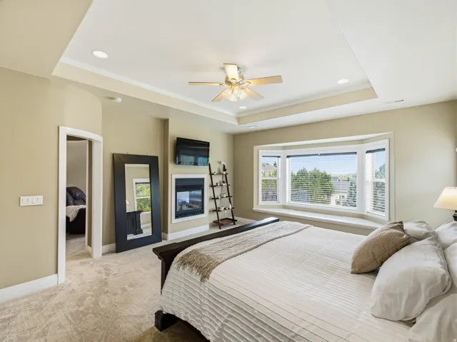 Spacious room featuring a tiered tray ceiling with recessed lighting, a ceiling fan, plush carpeting, a built-in fireplace, and a large bay window with a deep sill