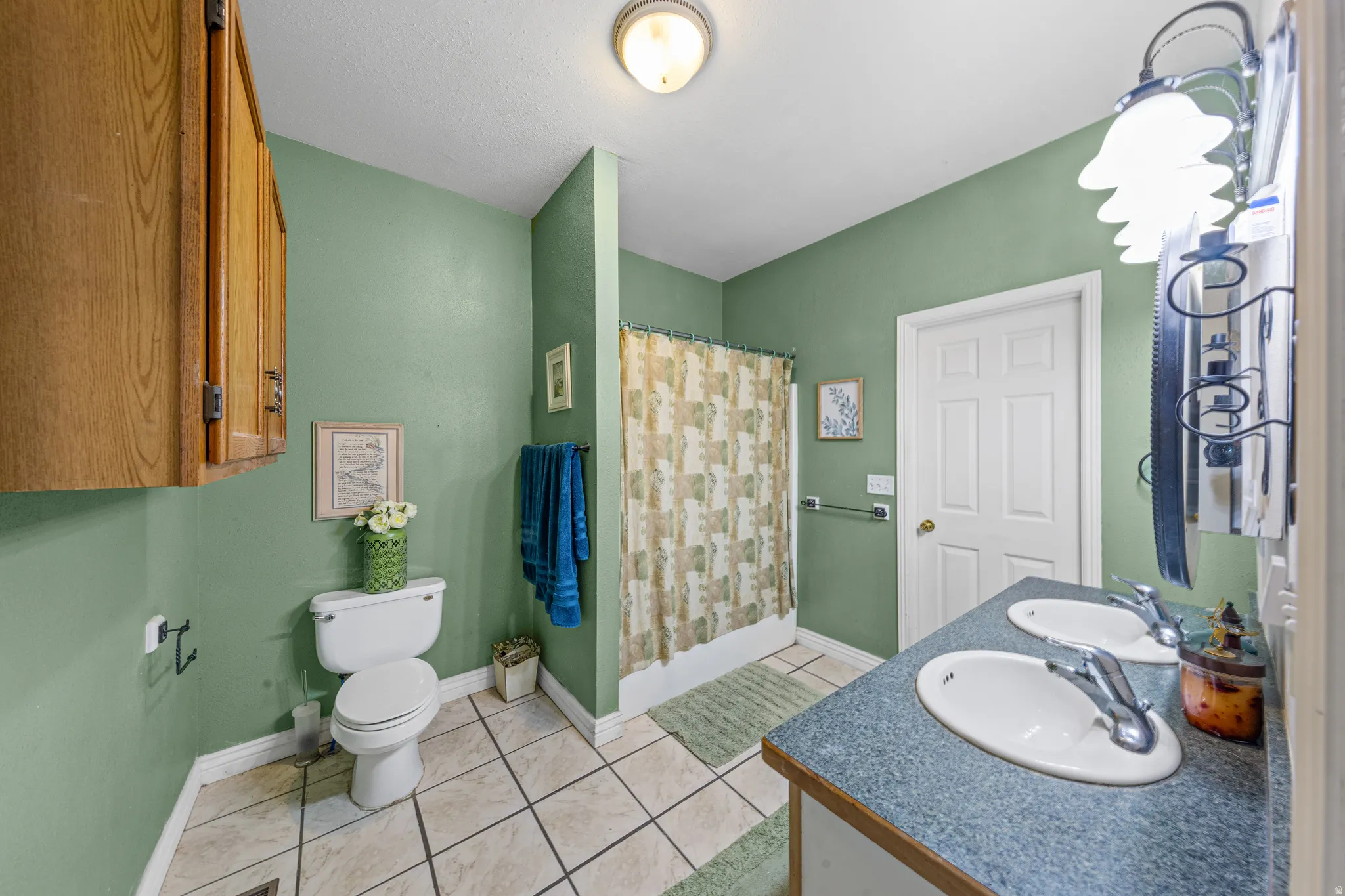 Bathroom featuring a dual-sink vanity with a blue countertop, a separate toilet area, a shower with a patterned curtain, overhead lighting, and wood-finish cabinetry