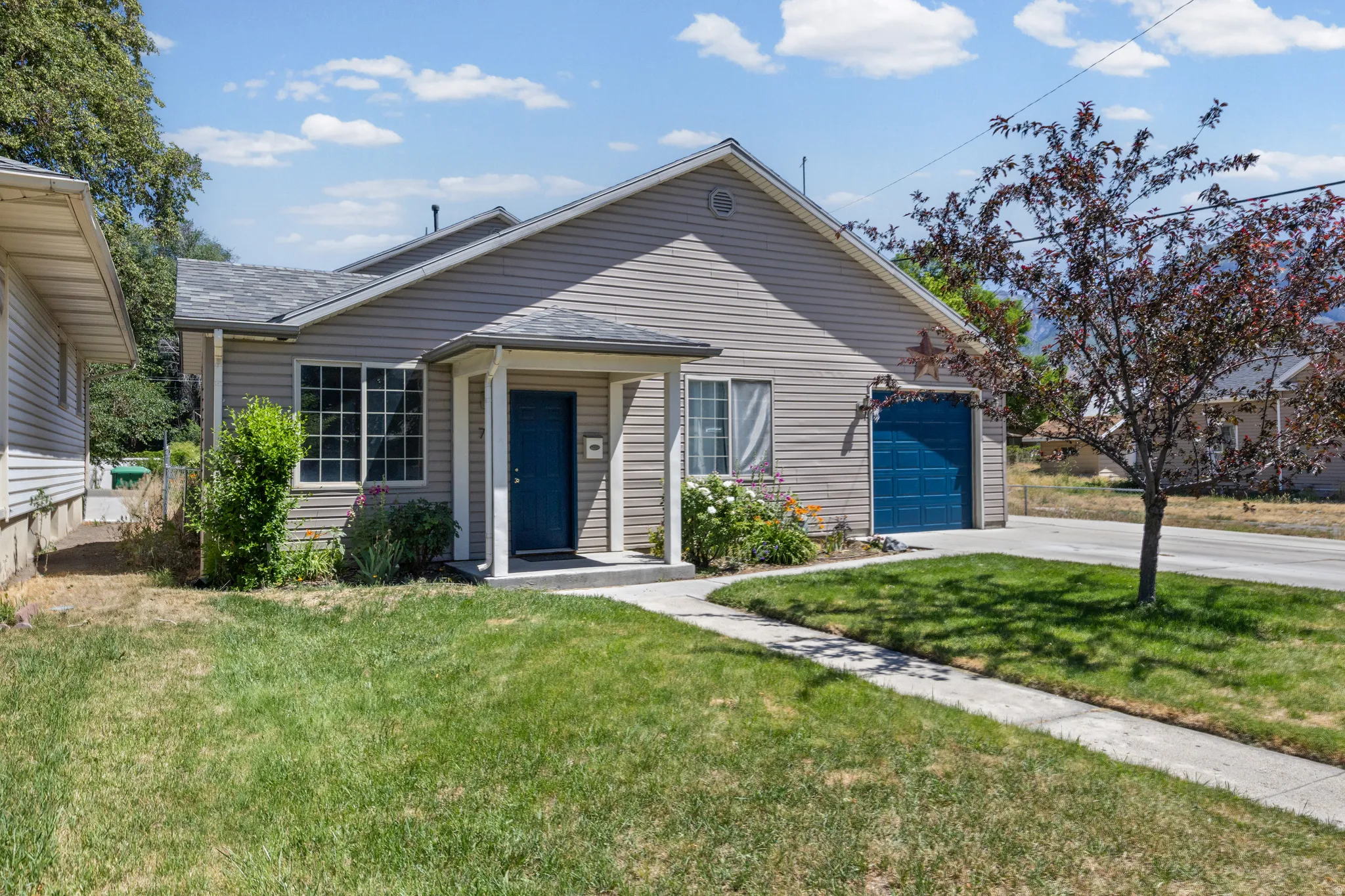 Single-story residence featuring light gray siding, a covered entry porch, a blue front door, a blue garage door, and an established lawn with a concrete sidewalk