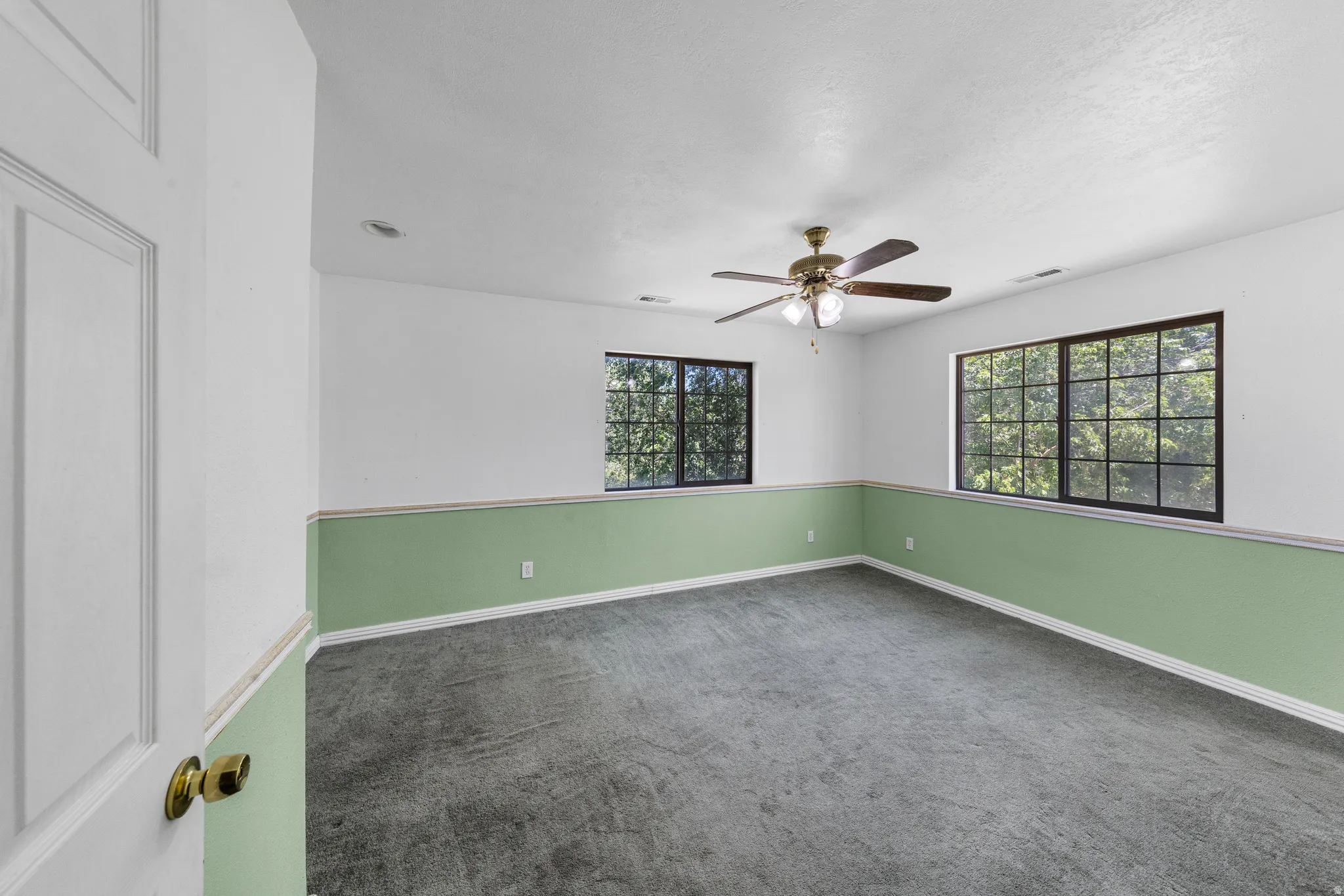 Spacious room featuring multiple large windows with black grilles, a brass-finish ceiling fan with integrated lighting, and carpet flooring