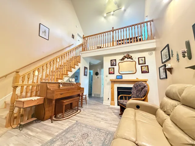 Living area with vaulted ceilings, wood-finish flooring, a prominent wooden staircase with balusters, a built-in fireplace with a white brick surround and wood mantel, and an upper-level balcony with a matching wooden railing