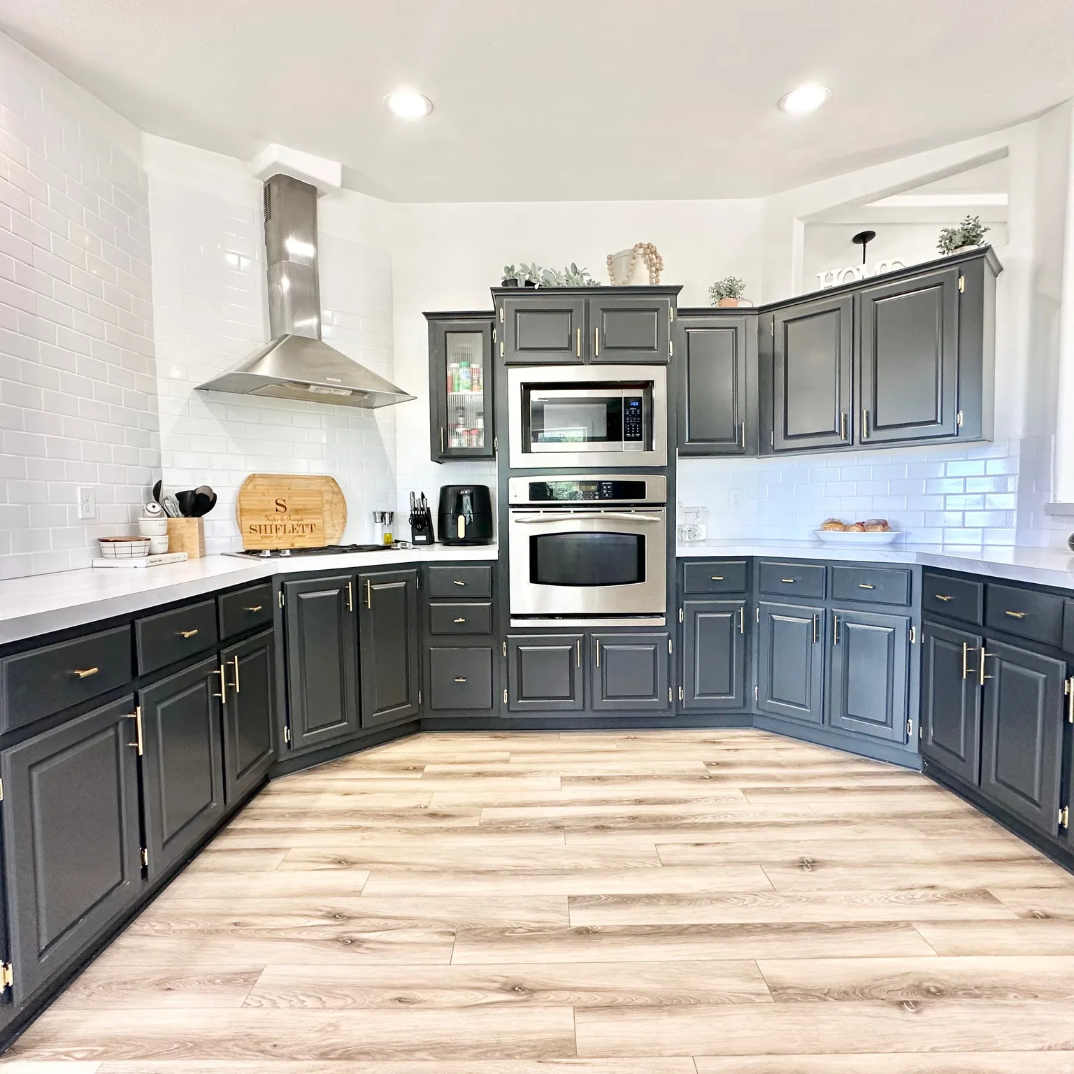 Kitchen featuring dark-finish cabinetry with gold-tone hardware, white subway tile backsplash, light-tone countertops, stainless steel appliances, and wood-finish flooring