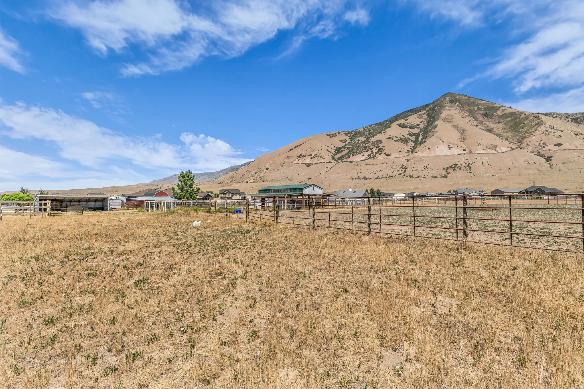 Expansive open field with perimeter fencing, featuring a barn structure, additional outbuildings, and mountain scenery